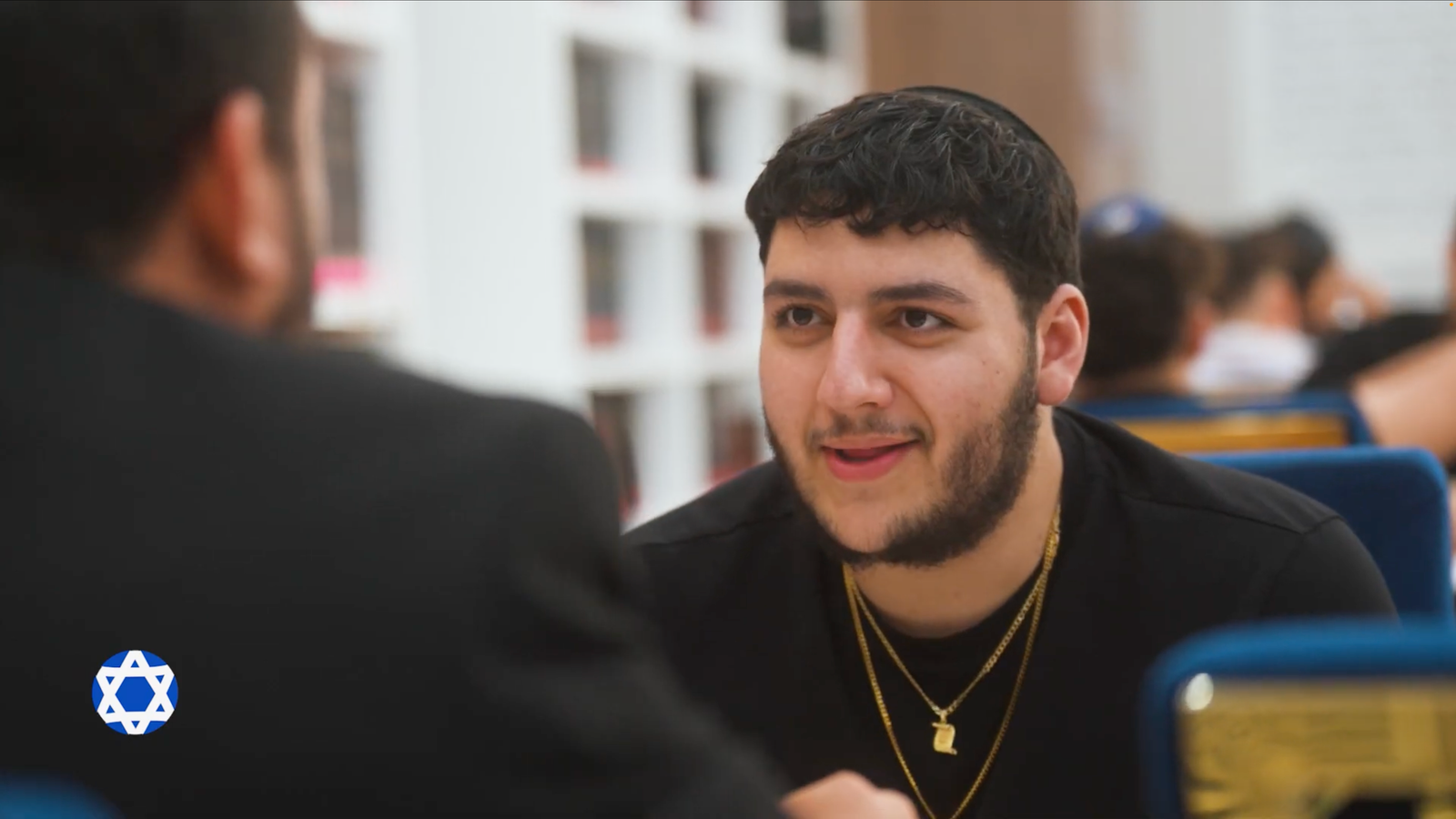 Young man in a black shirt and gold chains talking across a table in a bright hall
